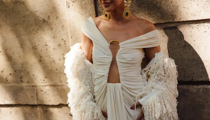 Elegancka sesja fashion: modelka w kremowej sukni z wycięciem i futrzanym bolerkiem, ozdobiona złotą biżuterią.