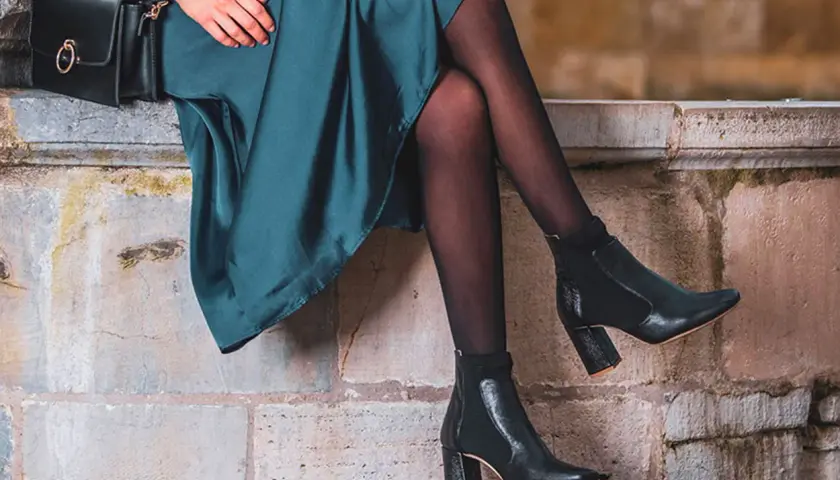 Jambes élancées en collant noir, chaussées de bottines à talon. Une robe vert émeraude et un sac à main complètent ce look chic.