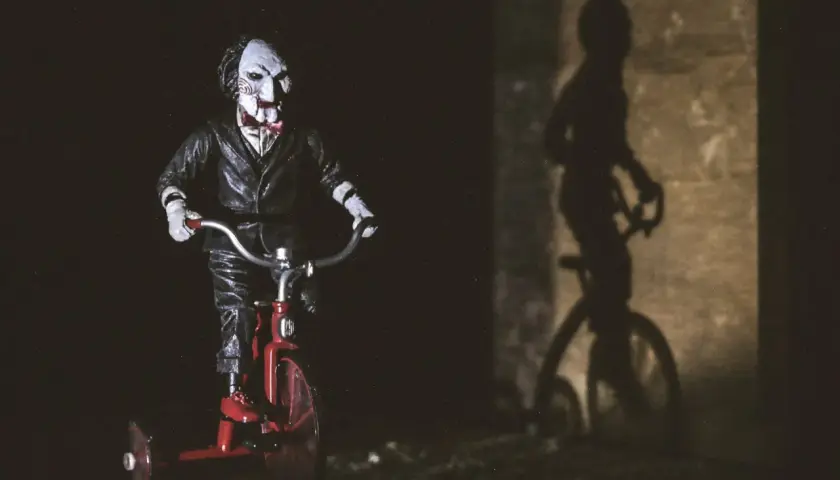 Lalka Billy z Piły na czerwonym trójkołowcu, przypominająca scenę z najlepszych horrorów. Cień na ścianie dodaje grozy.