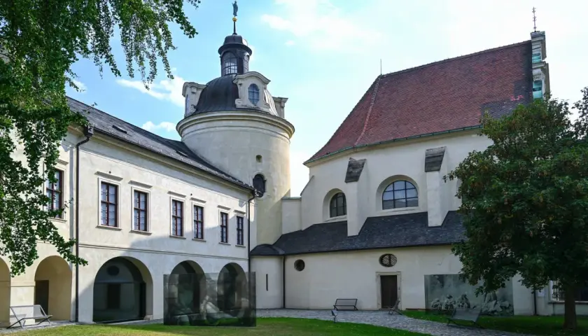 Ołomuniec zamek: okrągła wieża z kopułą i figurą na szczycie, obok budynek z arkadami i kościół z czerwoną dachówką.