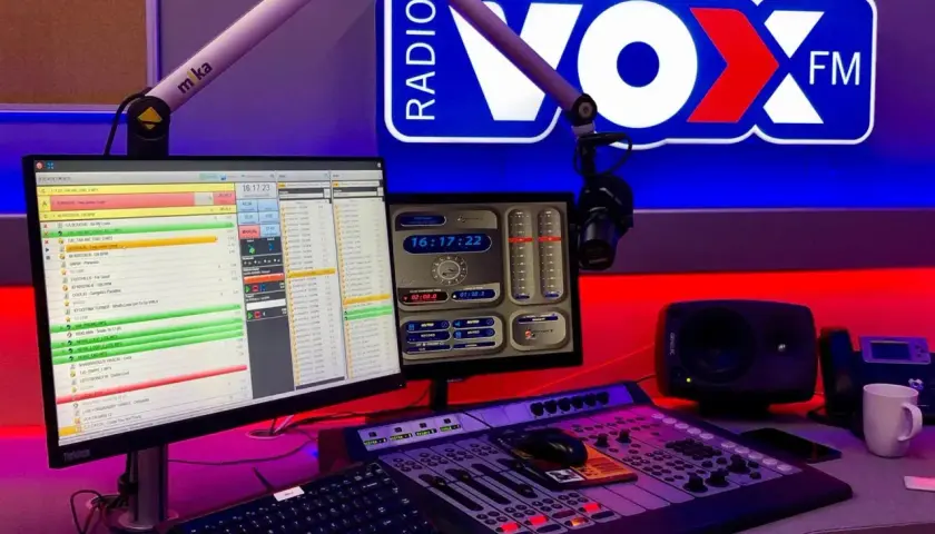 Studio radiowe VOX FM Warszawa. Komputery z listą utworów, mikser, mikrofon i logo stacji.