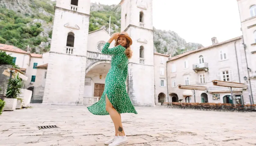 Une femme porte une robe verte fleurie et des baskets blanches, un chapeau de paille sur la tête, dans une place pavée devant une église.
