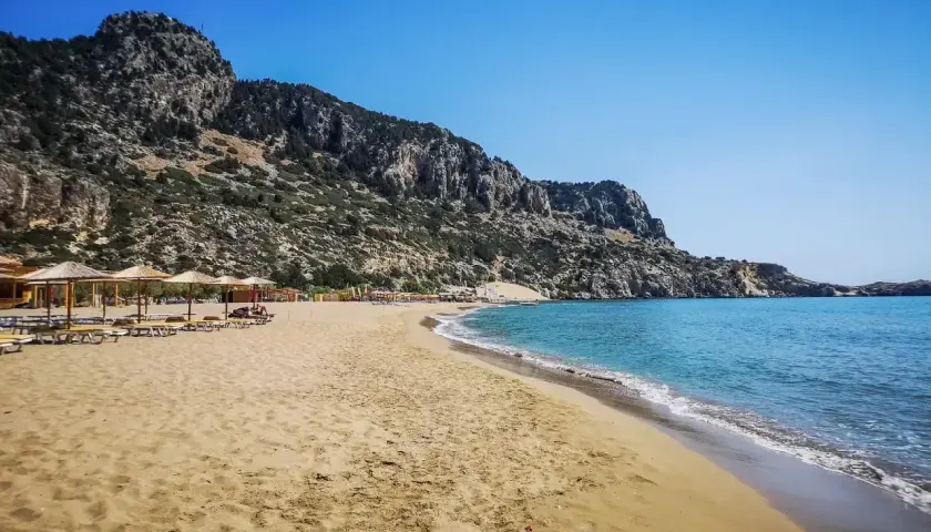 Piękna, piaszczysta plaża Tsambika z turkusową wodą i skalistym wzgórzem w tle. Leżaki i parasole czekają na plażowiczów.
