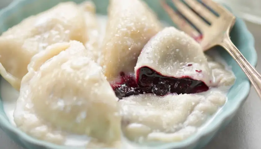 Pyszne pierogi z jagodami, polane śmietaną i posypane cukrem, czekają na widelca.