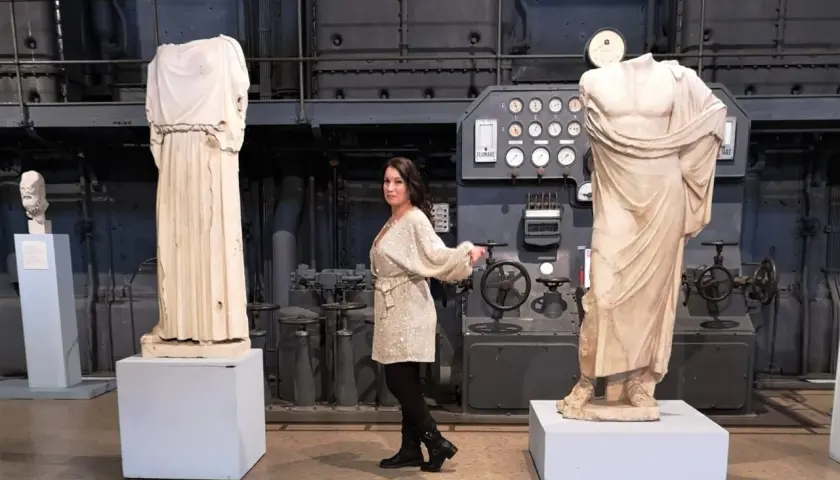 Donna in abito scintillante tra statue classiche e macchinari industriali, forse nel vecchio gasometro.