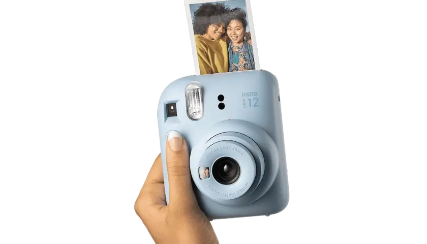 Niebieski aparat Instax Mini 12, trzymany w dłoni, z właśnie wysuniętym, jasnym zdjęciem dwóch uśmiechniętych przyjaciółek.