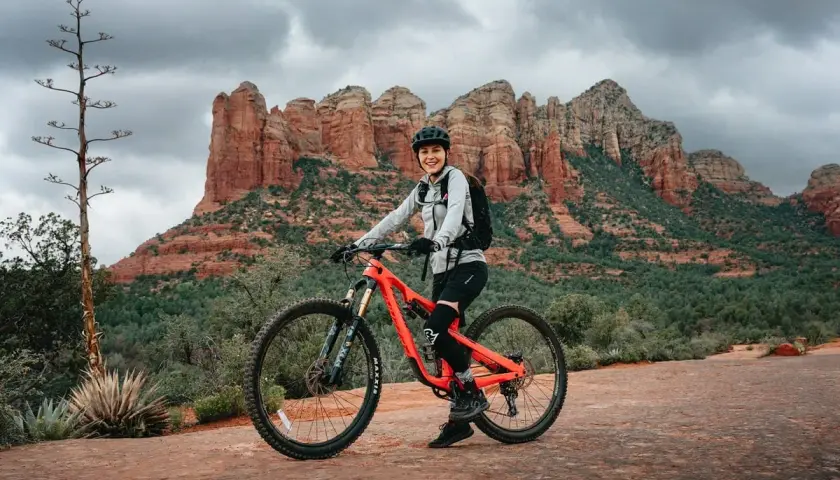 A smiling woman in riding gear poses on her bright orange mountain bike, ready for a trail tune-up amidst the stunning red rock formations of Sedona.