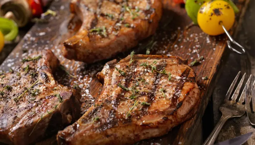 Trois côtelettes de porc grillées, garnies d'herbes et de gros sel, sur une planche en bois. Une surprise de porc barbecue prête à être dégustée.
