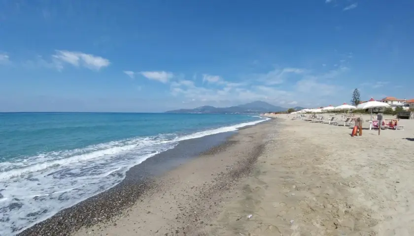 Plage de sable et de galets à Ascea Marina, avec des vagues bleues, des parasols blancs et des montagnes à l'horizon.