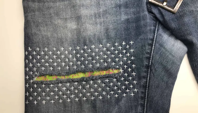 Close-up of denim jeans with a colorful fabric patch and white cross-stitch embroidery, showcasing a stylish sashiko jeans repair.