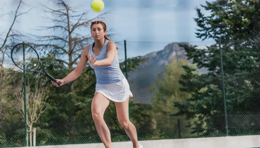 Kobieta w białej spódniczce i niebieskiej koszulce ćwiczy tenis przy ścianie, odbijając piłkę rakietą.