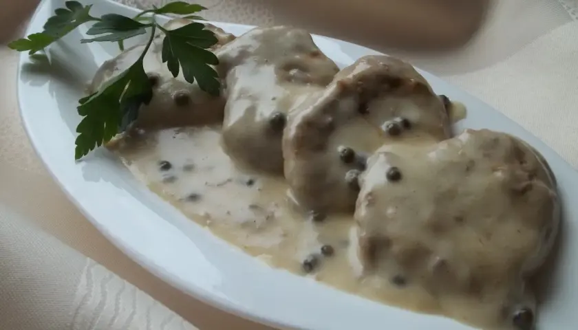 Polędwiczki w sosie z zielonego pieprzu, udekorowane natką pietruszki, prezentują się apetycznie na białej, łódkowatej zastawie.