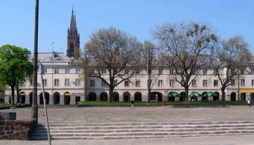 Łódź rynek z kamienicami, drzewami i wieżą kościelną w tle.