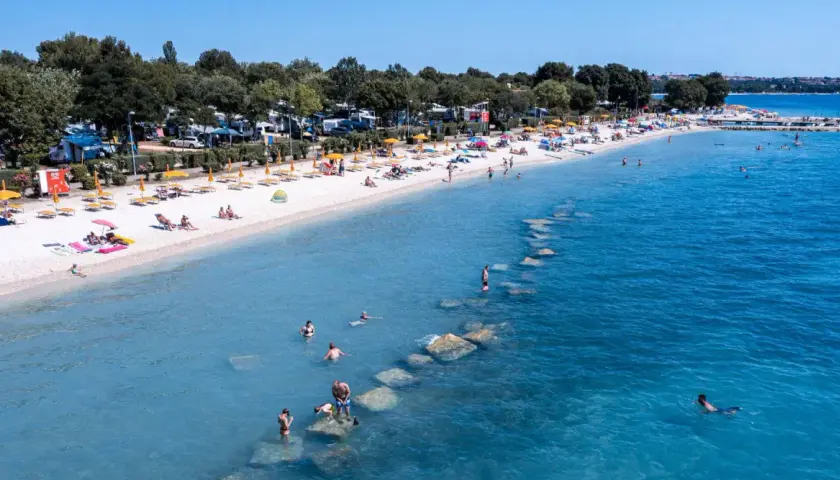 Słoneczna plaża z turystami, parasolami i kamperami w tle. Woda kusi do kąpieli, a kamienie tworzą naturalne falochrony. Idealne miejsce na bi village camping.