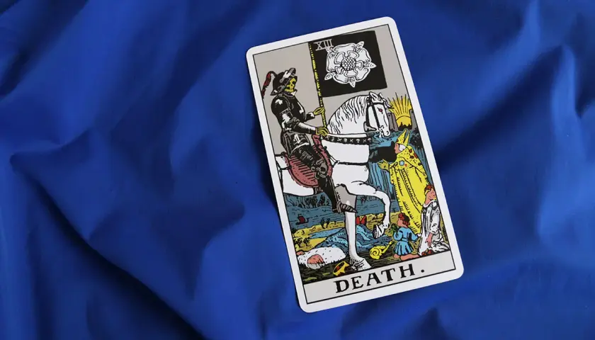 Tarot karta Śmierci na niebieskim tle. Postać w zbroi na białym koniu, niosąca flagę z różą, symbolizuje nieuchronne zmiany i transformację.