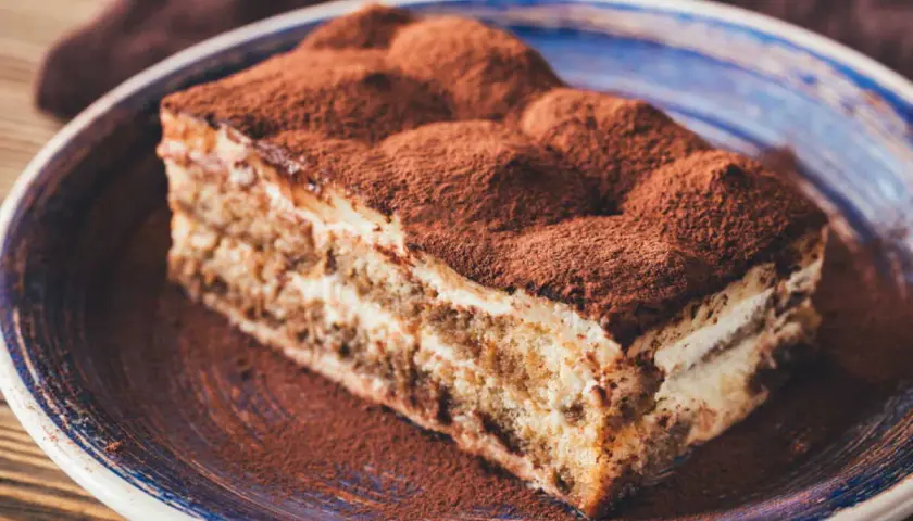Une délicieuse part de tiramisu, généreusement saupoudrée de cacao, sur une assiette bleue.