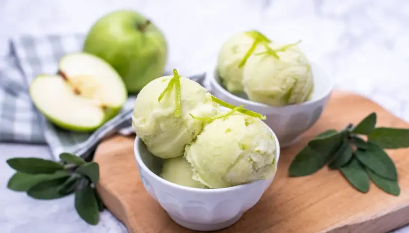 Orzeźwiający sorbet jabłkowy w miseczkach, udekorowany skórką z limonki. Obok całe i połówka zielonego jabłka.