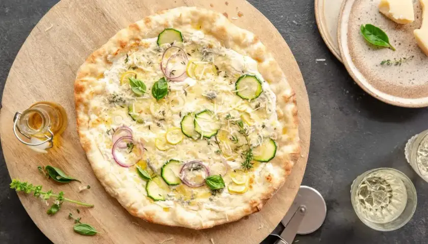 Apetyczna pizza bianca z cukinią, cebulą i ziołami na drewnianej desce. Obok buteleczka oliwy i kieliszki z winem.