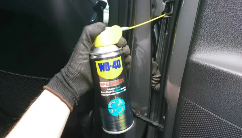 Ręka w rękawiczce aplikuje biały smar litowy WD-40 na zawias drzwi samochodu.