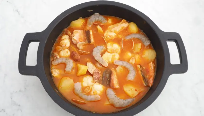 Un delicioso guiso marinero burbujea en una cazuela negra, con patatas, gambas, pescado y almejas en una salsa rojiza.