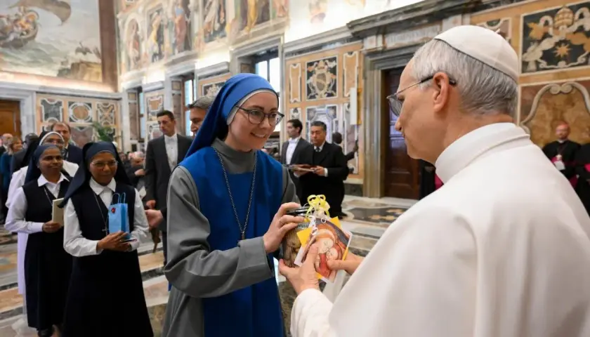 Eine Ordensfrau überreicht dem Papst ein Geschenk. Im Hintergrund stehen weitere Ordensschwestern und Männer in Anzügen.