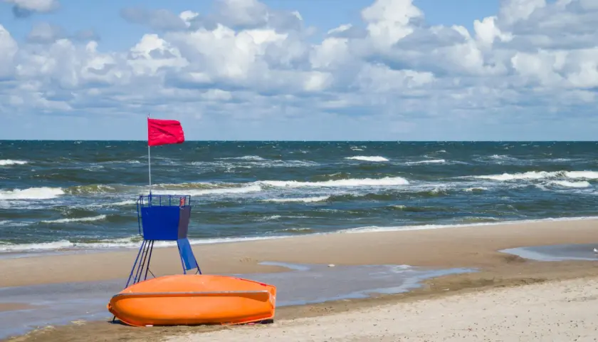 Czerwona flaga na wieży ratowniczej, pomarańczowa łódź ratownicza na plaży, wzburzone morze.