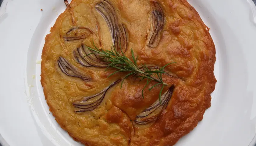 Herzhafte Kichererbsenmehl Rezepte: Goldbrauner Kuchen mit roten Zwiebeln und Rosmarin auf weißem Teller.