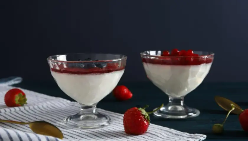 Dwa pucharki z panna cottą, udekorowane musem truskawkowym i owocami leśnymi. Obok leżą świeże truskawki i złota łyżeczka.