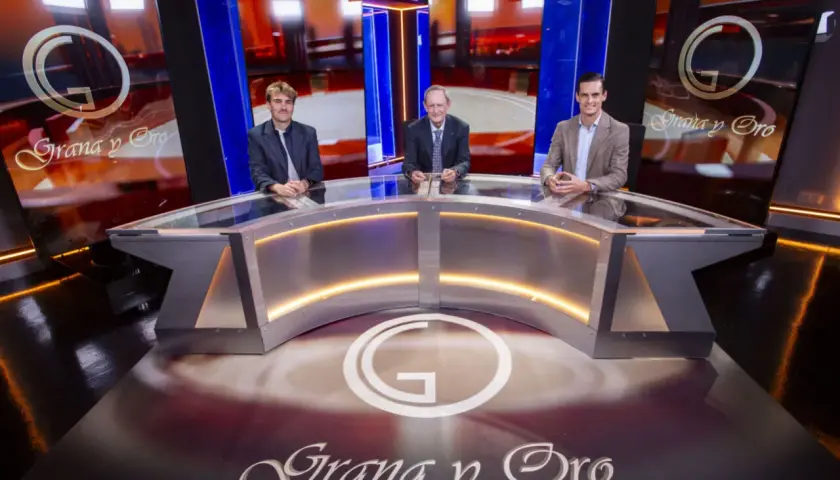 Tres hombres sentados en un plató de televisión con el logo "Grana y Oro" en el fondo y en el suelo.
