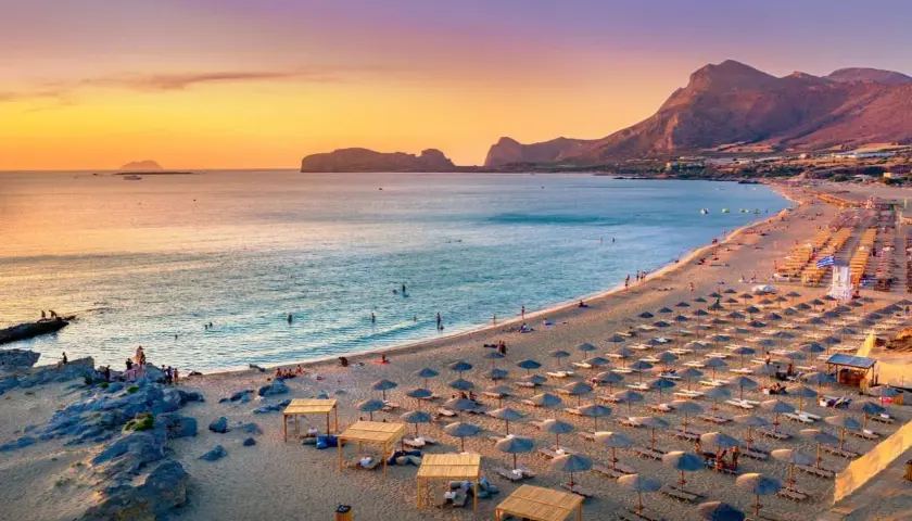 Ein langer Sandstrand mit vielen Liegestühlen und Sonnenschirmen. Das Meer ist ruhig, im Hintergrund Berge. Perfekt für die Frage: wo ist es im Mai warm Europa?