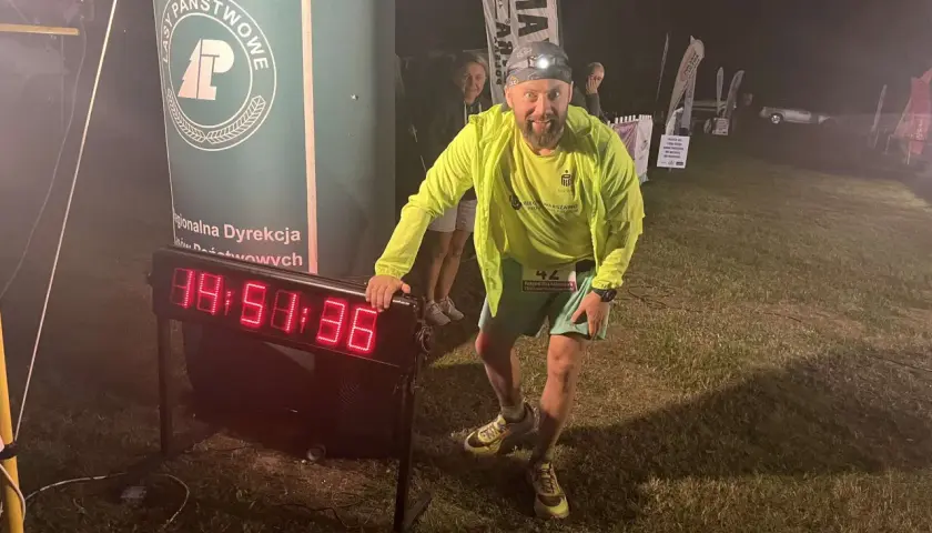 Uśmiechnięty biegacz z numerem 42 przy zegarze na trasie backyard ultra. Czołówka oświetla mu drogę.