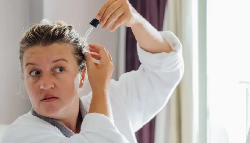 Kobieta w szlafroku aplikuje serum na skórę głowy. Wierzy, że goździki i rozmaryn na włosy przyniosą jej ulgę i wzmocnią kosmyki.