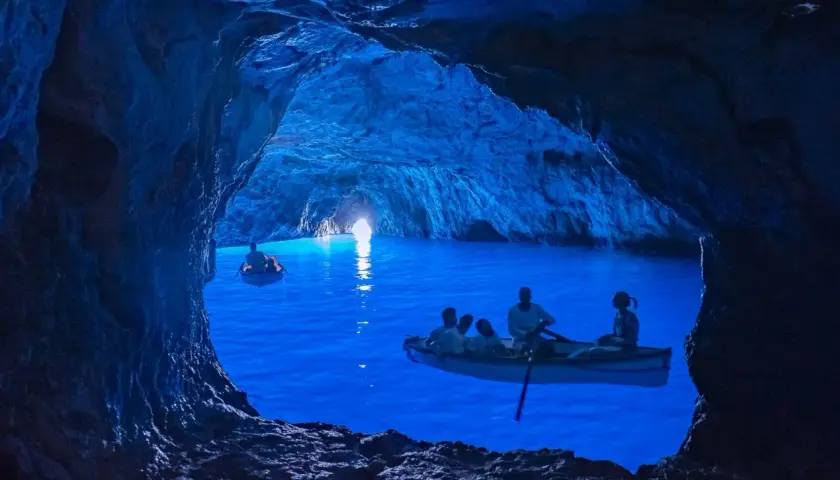 Barche a remi navigano nelle magiche grotte blu, illuminate da una luce surreale che rende l'acqua di un blu intenso.