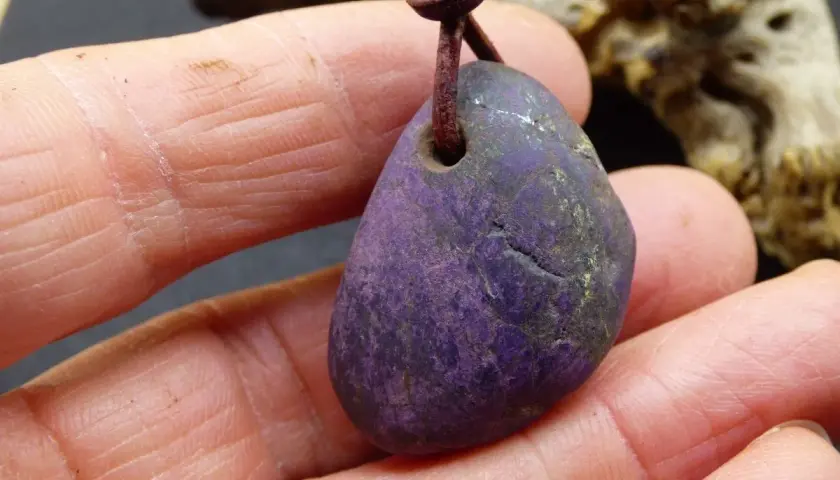 Ein purpurit Anhänger, gehalten in Fingern. Seine tiefviolette Farbe und die natürliche Textur lassen die purpurit wirkung erahnen.