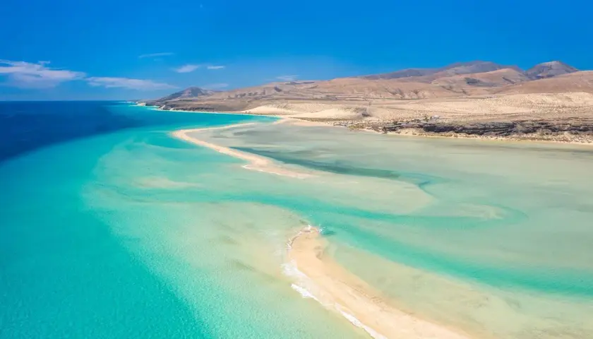 Aguas turquesas y arena dorada en Fuerteventura. Un paraíso natural para descubrir qué ver en Fuerteventura, con montañas áridas al fondo.