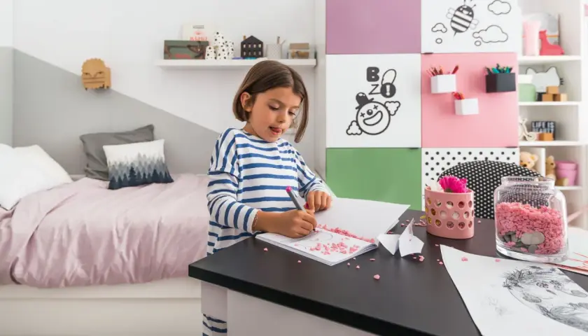 Dziewczynka rysuje w swoim małym pokoju dla dziecka. Na biurku leżą kolorowe kamyczki i papier.