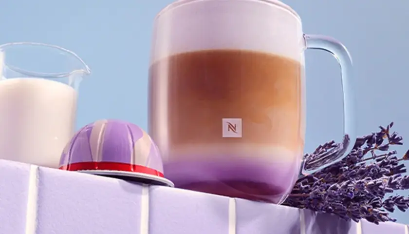 Ein aromatischer Lavendel Latte in einem Glasbecher, daneben eine Kaffeekapsel und Milch.