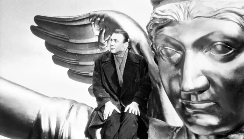 Un uomo seduto su un'ala d'angelo dorata, con un volto statuario di fronte. Un'immagine iconica per capire "il cielo sopra berlino".