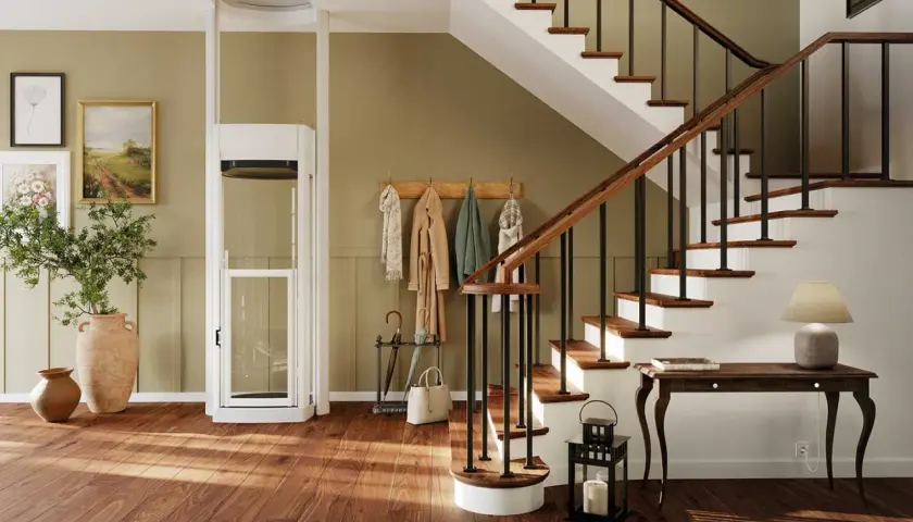 Ein moderner Treppenlift mit Glaskabine steht neben einer Holztreppe. Bilder und Garderobe schmücken die Wand.