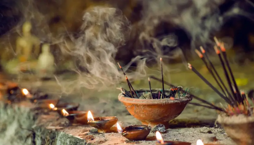 Humo de incienso llenando el aire, un recordatorio de que el incienso es malo para los pulmones. Velas encendidas y estatuillas borrosas en el fondo.