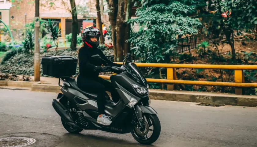 Mujer con casco y ropa negra conduce una moto Yamaha sin marchas, con caja trasera, por una calle arbolada.