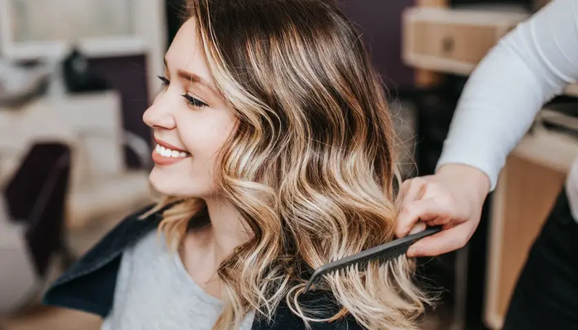 Sonrisa radiante con un balayage rubio natural, el cabello ondulado y con mechas doradas resalta su belleza.