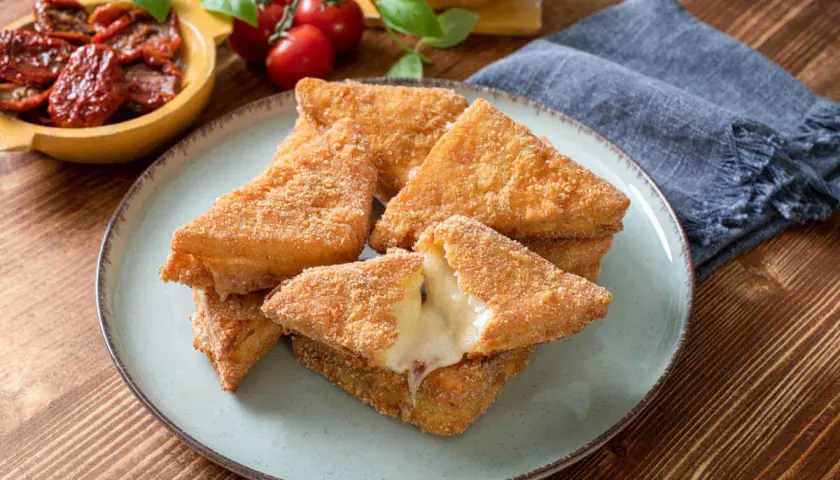 Chrupiące mozzarella in carrozza, z rozpływającym się serem w środku, na talerzu. Obok pomidorki i suszone pomidory.