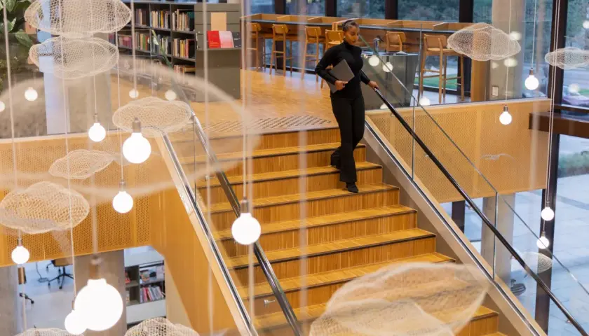 Kobieta z laptopem schodzi po drewnianych schodach w nowoczesnej bibliotece. Zastanawia się, ile trwają studia, gdy mija wiszące dekoracje w kształcie chmur.
