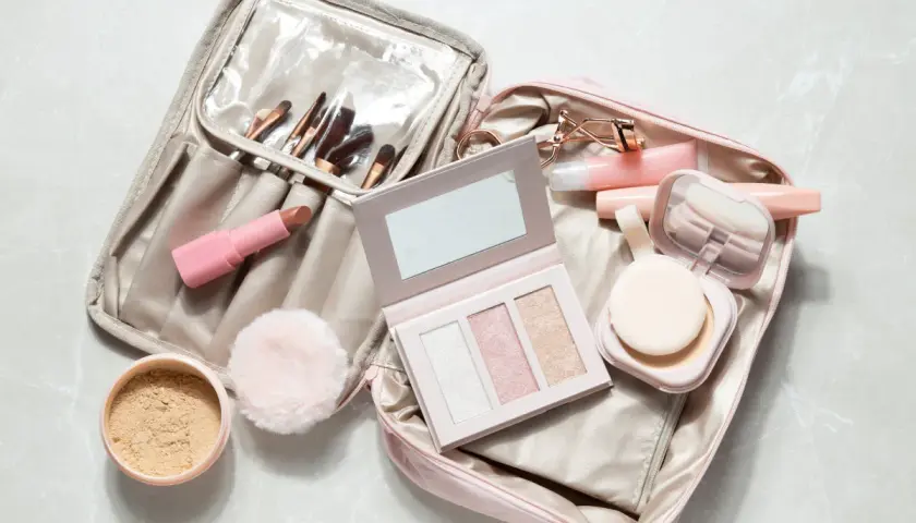 Une trousse de maquillage ouverte, prête pour un voyage en bagage cabine, révèle rouges à lèvres, pinceaux, palette et poudrier.
