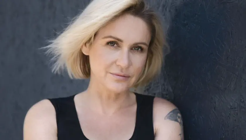 Małgorzata Ohme z blond włosami i tatuażem na ramieniu, na ciemnym tle.