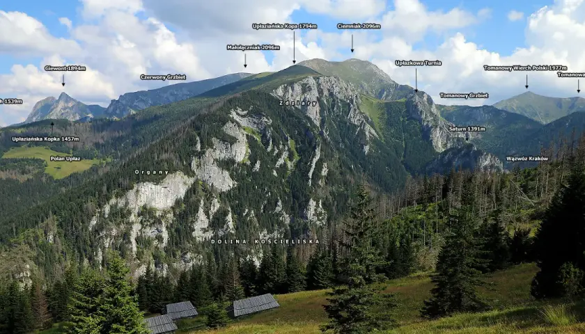 Widok na Tatry z Doliną Kościeliską. W oddali majaczą szczyty, a na pierwszym planie widać polanę z kilkoma drewnianymi halami stołami.