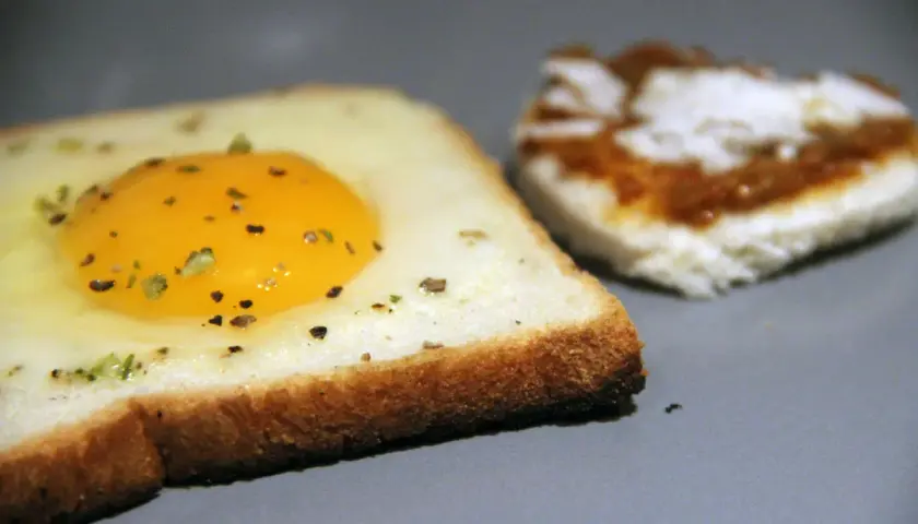 Une tranche de pain de mie avec un œuf au plat et des herbes. Une autre tranche est à côté, tartinée. Recette pain de mie œuf poêle facile.