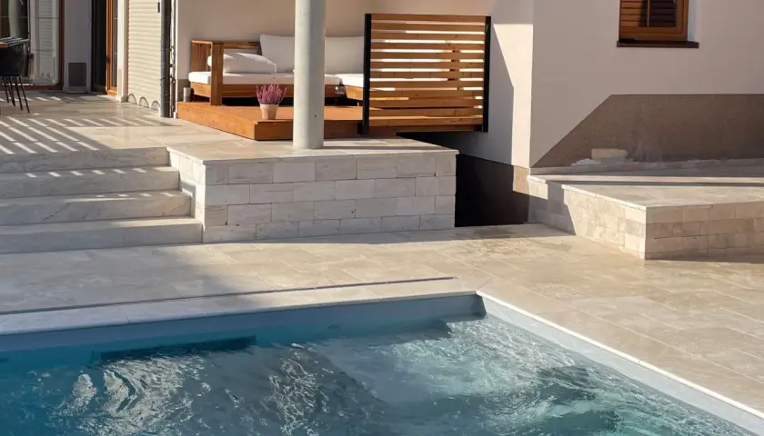 Piscina con bordo in pietra travertine. Gradini e pavimentazione in travertine creano un'elegante area relax con divani in legno.