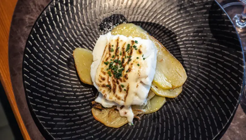 Bacalao gratinado con alioli, servido sobre patatas doradas en un plato negro texturizado.